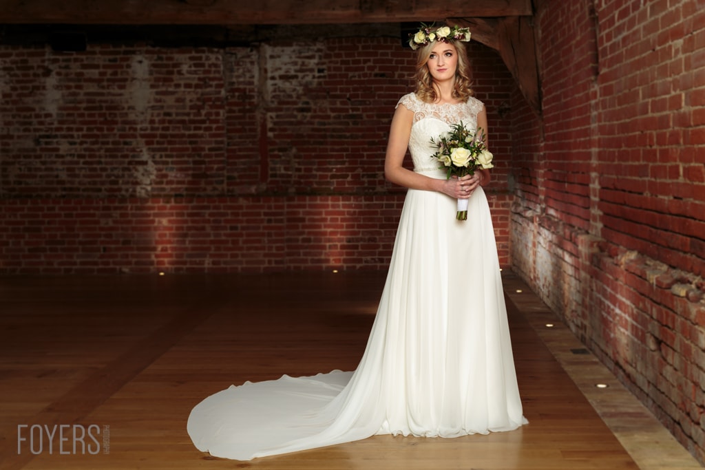 A bride in a white wedding dress and flower crown holds a bouquet, standing in an industrial brick corridor.