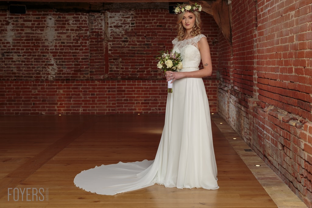 A bride in a white sleeveless lace and chiffon gown holds a bouquet in a brick-walled corridor.