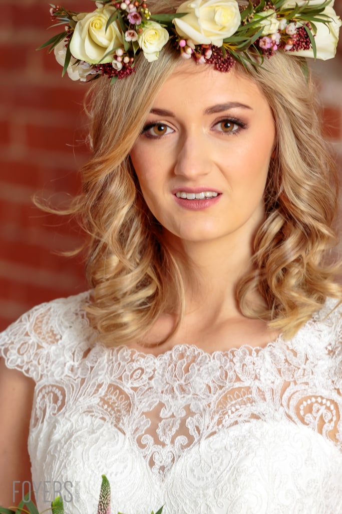 A woman with wavy blonde hair wearing a white lace top and a floral crown smiles at the camera against a red brick background.
