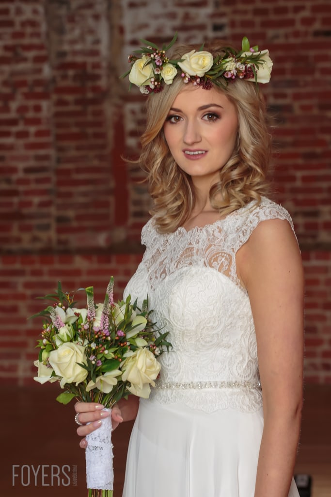 A bride in a white lace dress holds a bouquet of cream roses and burgundy flowers against a brick wall background.