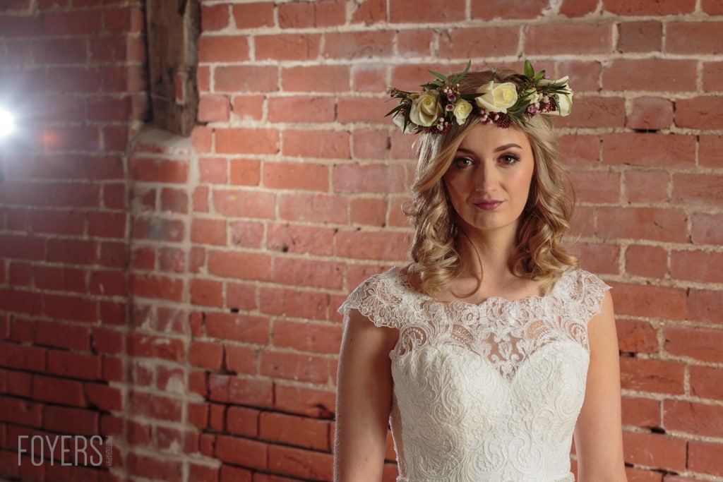 A woman in a white lace dress and flower crown stands against a red brick wall, looking at the camera.