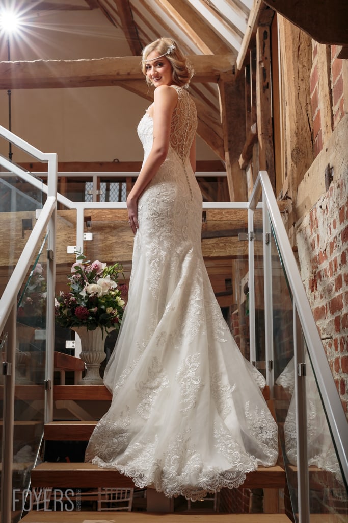 A bride in an elaborate white lace wedding dress with a train stands on wooden stairs in a rustic barn venue, smiling over her shoulder.