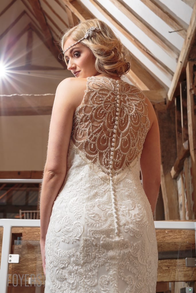 Woman in ornate white lace wedding dress viewed from behind, wearing beaded headband, in modern interior with wooden beams.