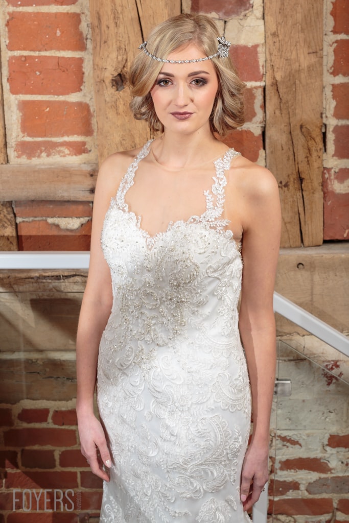 A woman wearing an ornate white beaded wedding dress and decorative headpiece stands against a rustic brick and wood backdrop.