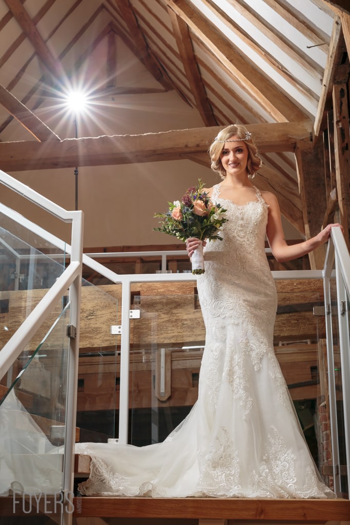 A bride in a white lace wedding dress holds a bouquet while standing in a barn venue with exposed wooden beams.