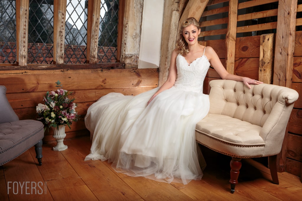 A bride in a white tulle wedding dress sits on a beige armchair in a rustic wooden interior, smiling at the camera.