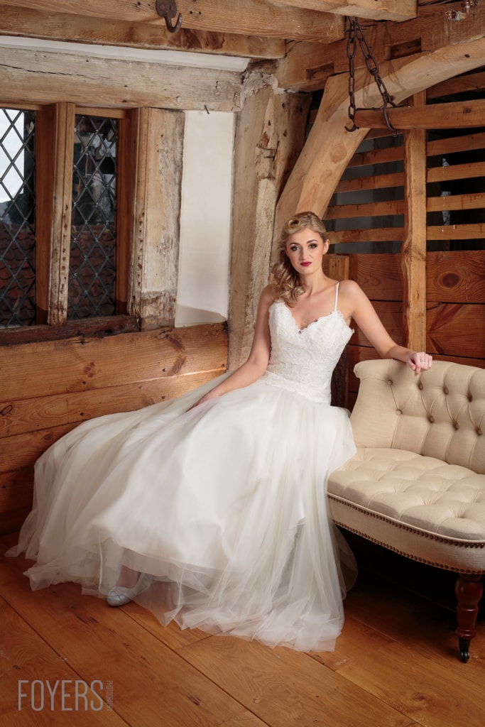A woman in a white wedding dress sits on a cream-coloured chair inside a rustic wooden barn or cottage.