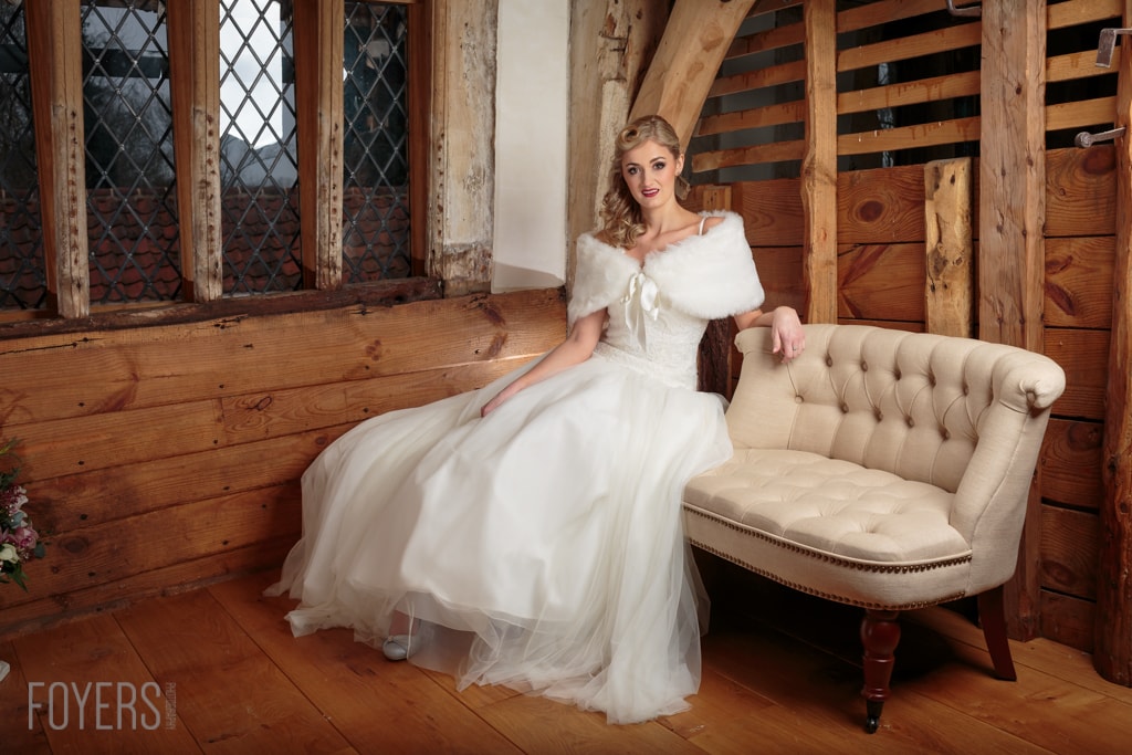 A woman in a white wedding dress with a shawl sits on a beige armchair in a wooden barn interior.