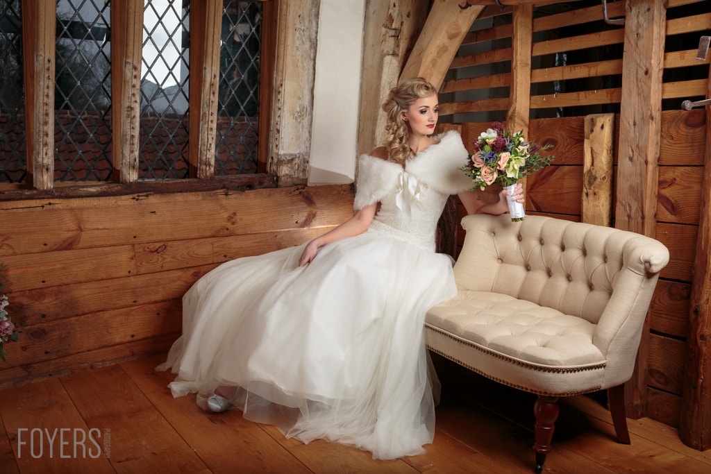 A bride in a white gown with fur stole sits on a beige armchair, holding a bouquet, in a wooden barn setting.