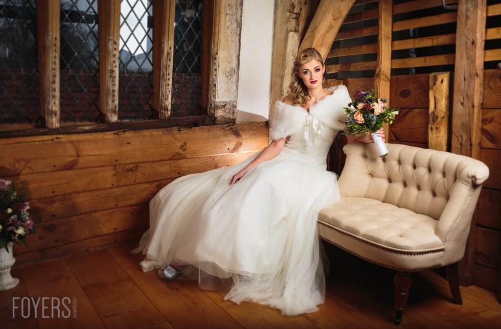 A bride in a white wedding dress with a fur stole sits on a cream armchair, holding a bouquet in a timber-framed room.