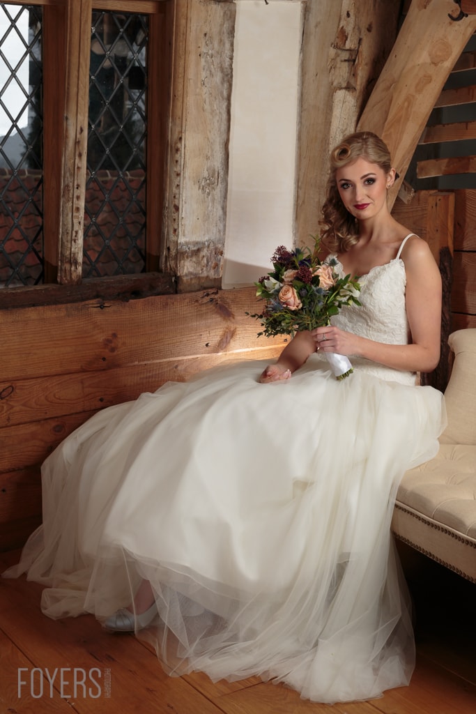 A bride in a white tulle gown sits holding a bouquet of flowers in a rustic wooden interior.