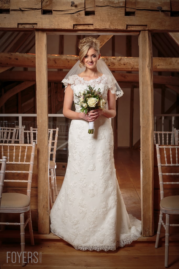 A bride in a white lace wedding dress and veil holds a bouquet, standing in a wooden barn venue with white chairs.