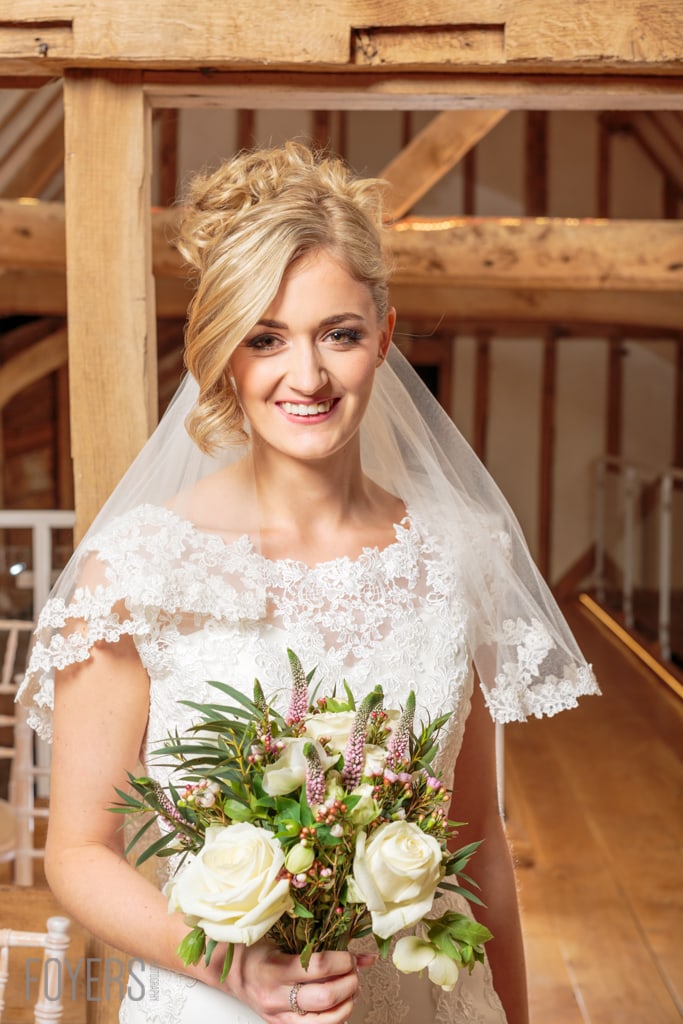 A smiling bride in a white lace dress and veil holds a bouquet of white roses and pink flowers in a timber-framed building.