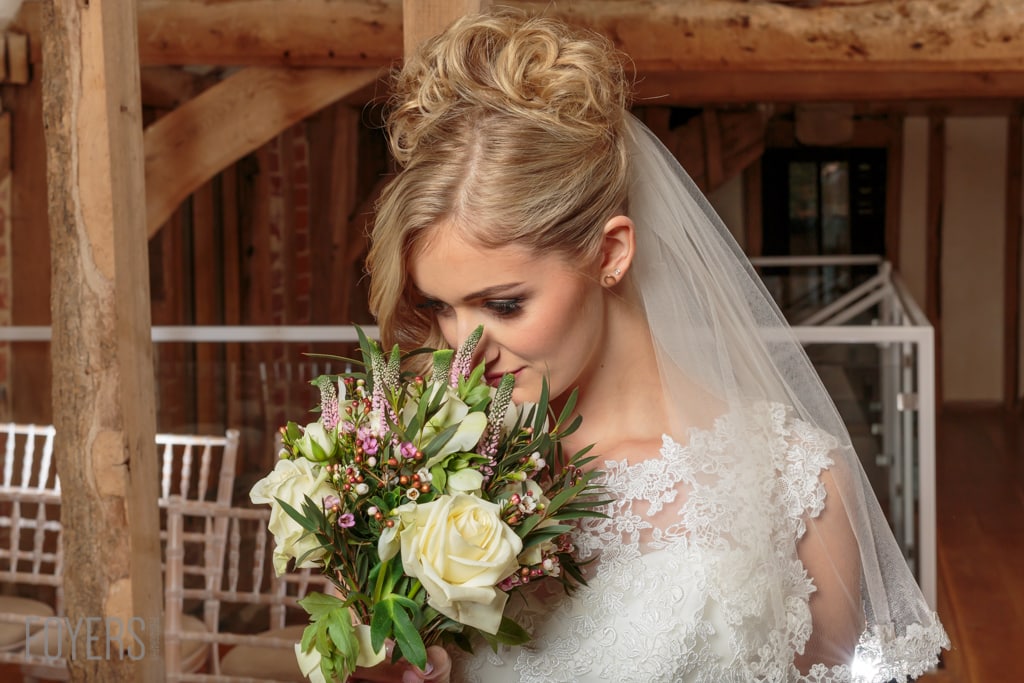 A bride in a white lace dress and veil holds a bouquet of cream roses and pink flowers in a rustic barn setting.