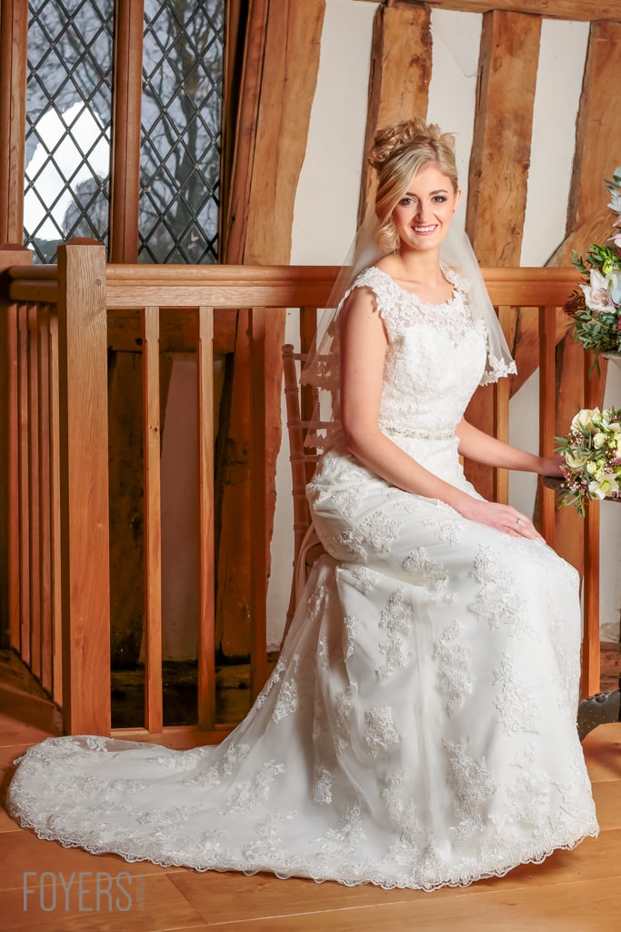 Bride in white lace wedding dress and veil sitting on wooden bench in timber-framed room with latticed windows.