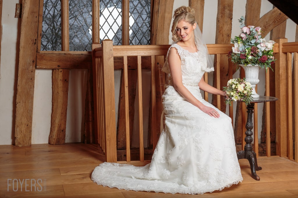 A bride in a white lace wedding dress and veil sits on a wooden bench in a timber-framed church interior.