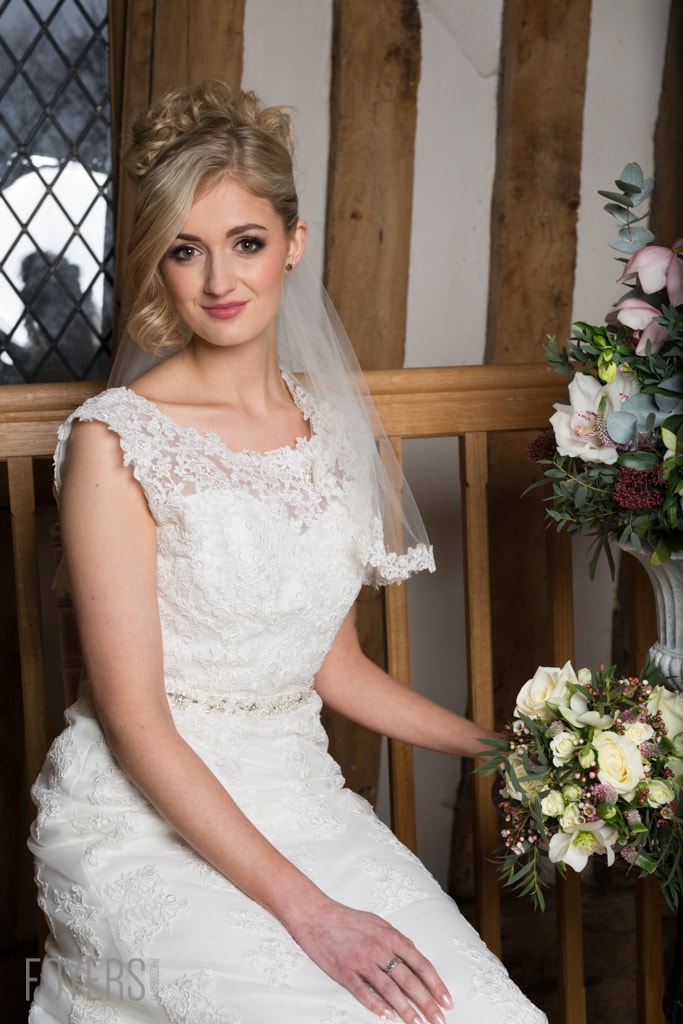 A bride in a white lace wedding dress and veil sits holding a bouquet of pink and white flowers in a timber-framed room.