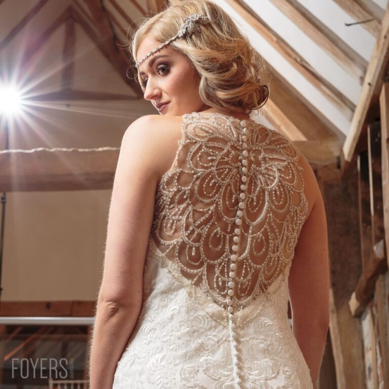 A woman wearing an ornate beaded wedding dress with decorative back detail, viewed from behind. She wears a beaded headpiece.