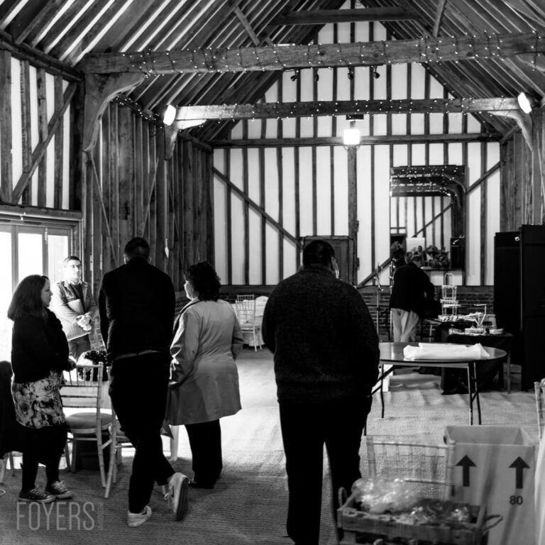 Black and white photograph of several people standing in a timber-framed barn or hall with vertical striped walls and exposed beams.