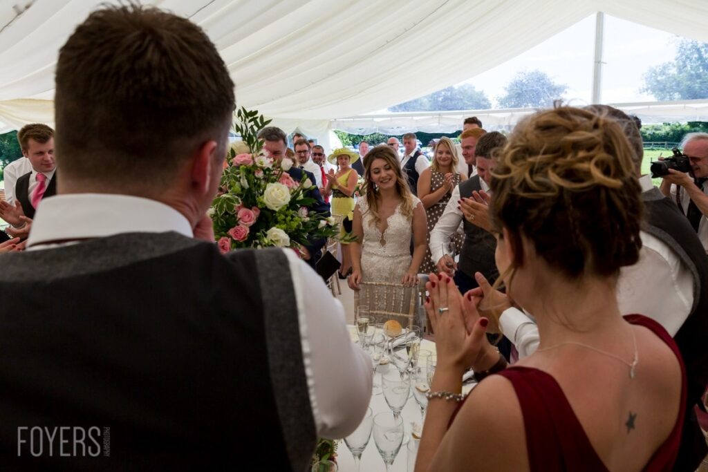 A bride in a white lace dress stands at a table under a marquee during a wedding reception, surrounded by guests.