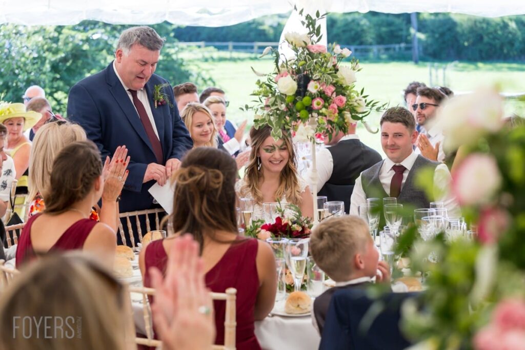 A man in a dark suit gives a speech at an outdoor wedding reception table, with guests seated around listening.
