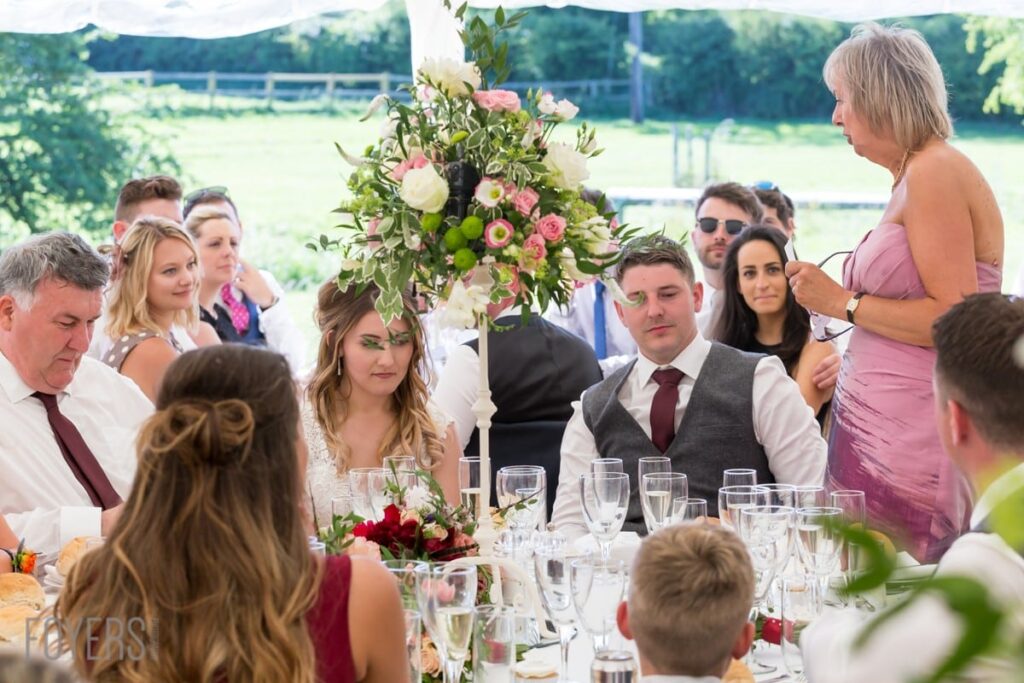 Wedding reception outdoors under a marquee. A woman in a pink dress stands speaking at the table while guests listen. Floral centrepiece visible.