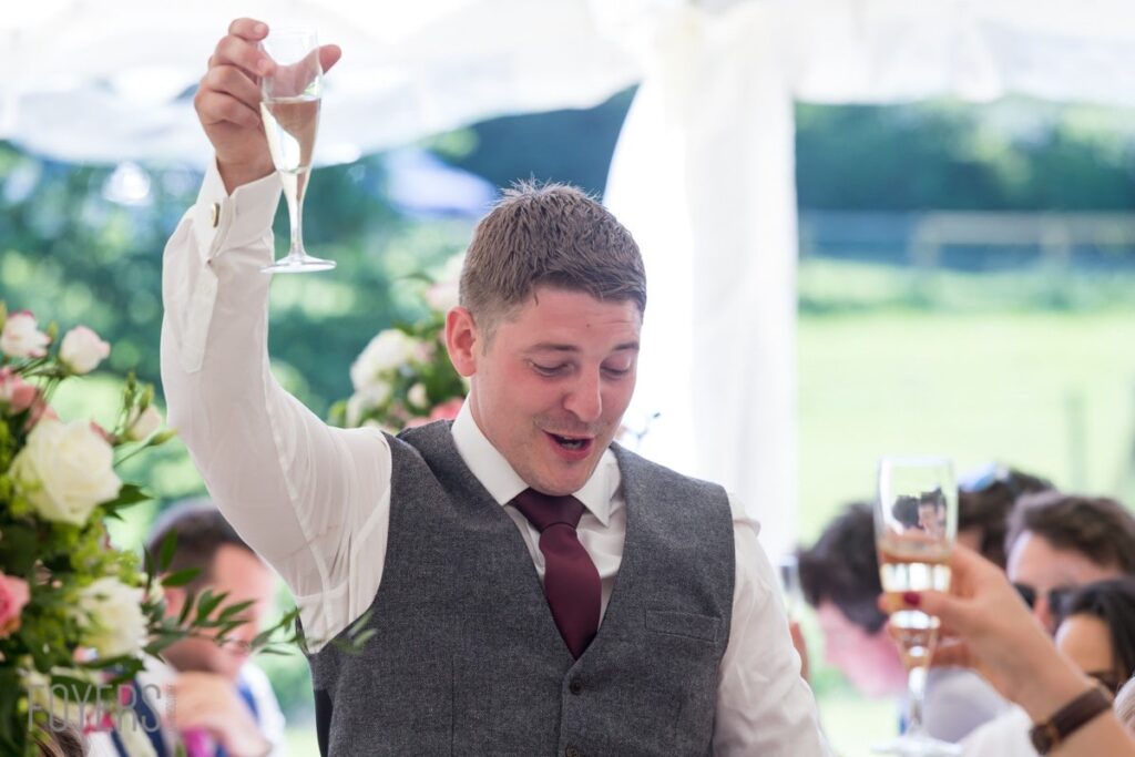 A man in a grey waistcoat and burgundy tie raises a champagne glass at an outdoor wedding reception.