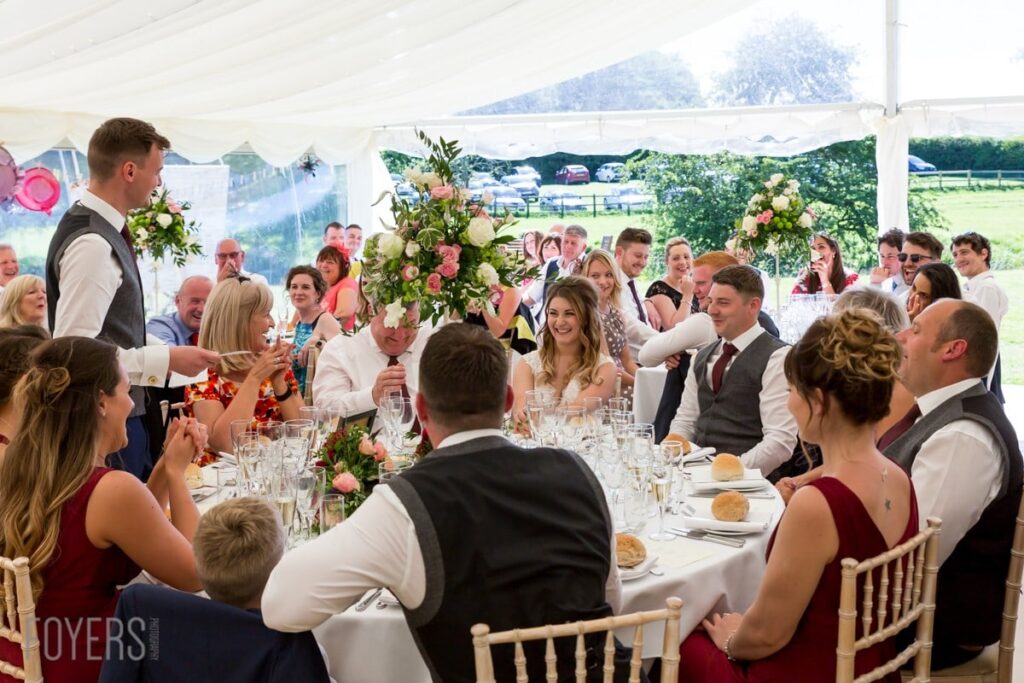 Wedding reception under a marquee with guests seated at tables, a man standing to give a speech, countryside visible beyond.