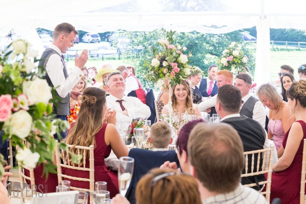 A man stands giving a speech at a wedding reception under a marquee, with guests seated at tables listening.