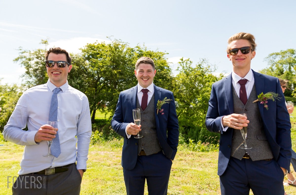 Three men in formal attire holding champagne glasses, standing outdoors in a garden setting on a sunny day.