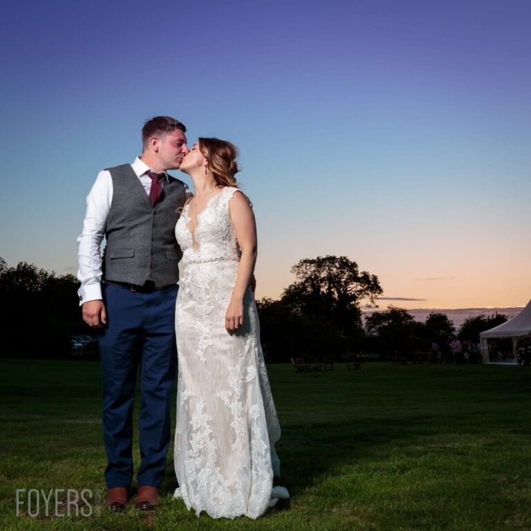A bride and groom kiss on a grassy field at dusk, with trees and a marquee visible in the background.