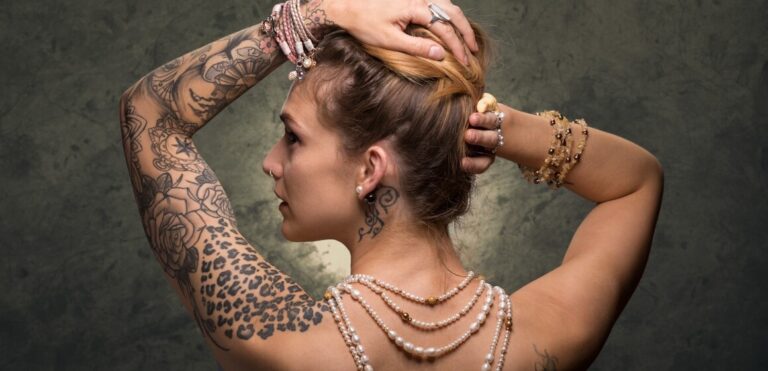 Woman with upswept hair and extensive tattoos on her arm, wearing pearl necklaces and rings, posed against a textured grey background.