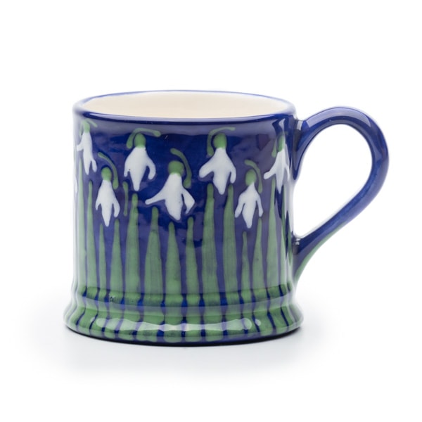 A blue ceramic mug with white snowdrop flowers and green stems painted around the top, shown against a white background.