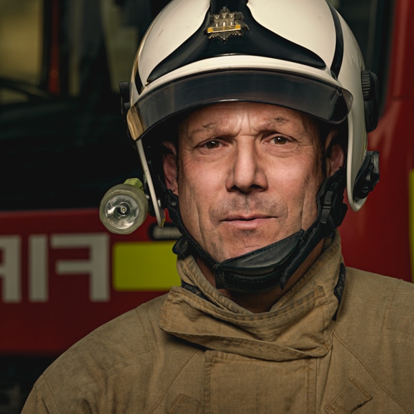 A firefighter wearing protective gear including a white and black helmet with a lamp, face shield, and tan jacket against a blurred fire station background.