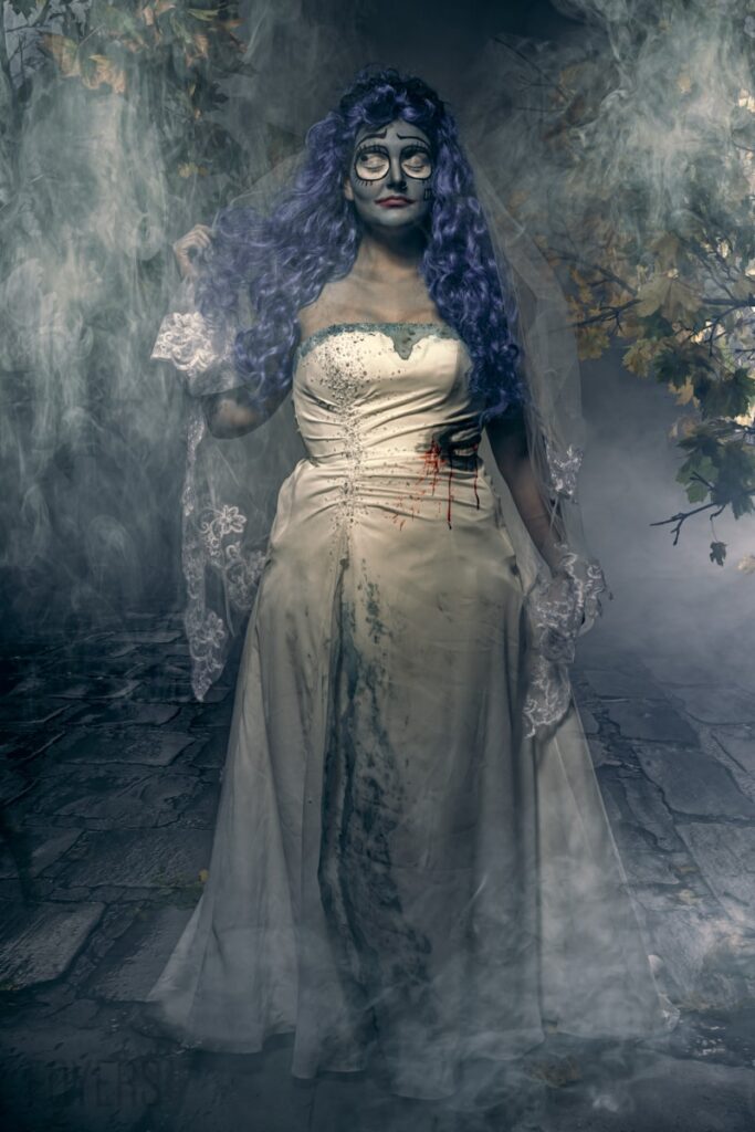 A woman with long purple hair and face paint wears a white wedding dress with red stains, standing in a misty, decaying stone setting.