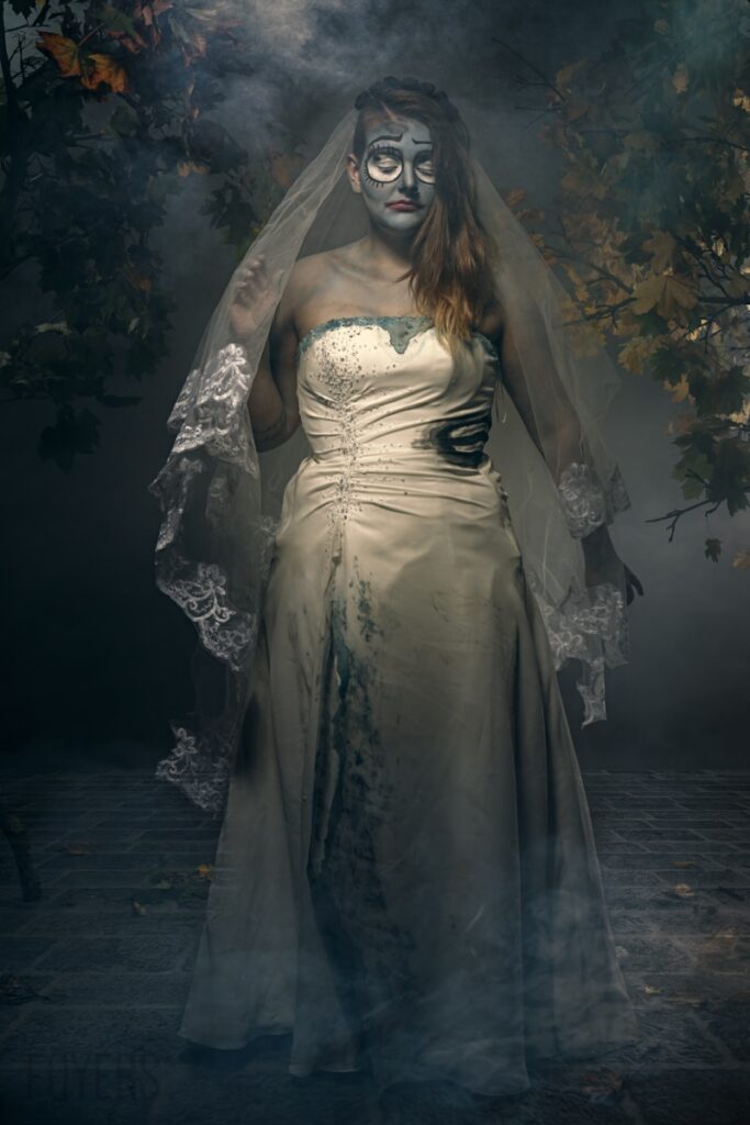 Woman in skull makeup and white wedding dress with lace veil stands in misty forest setting with autumn foliage.