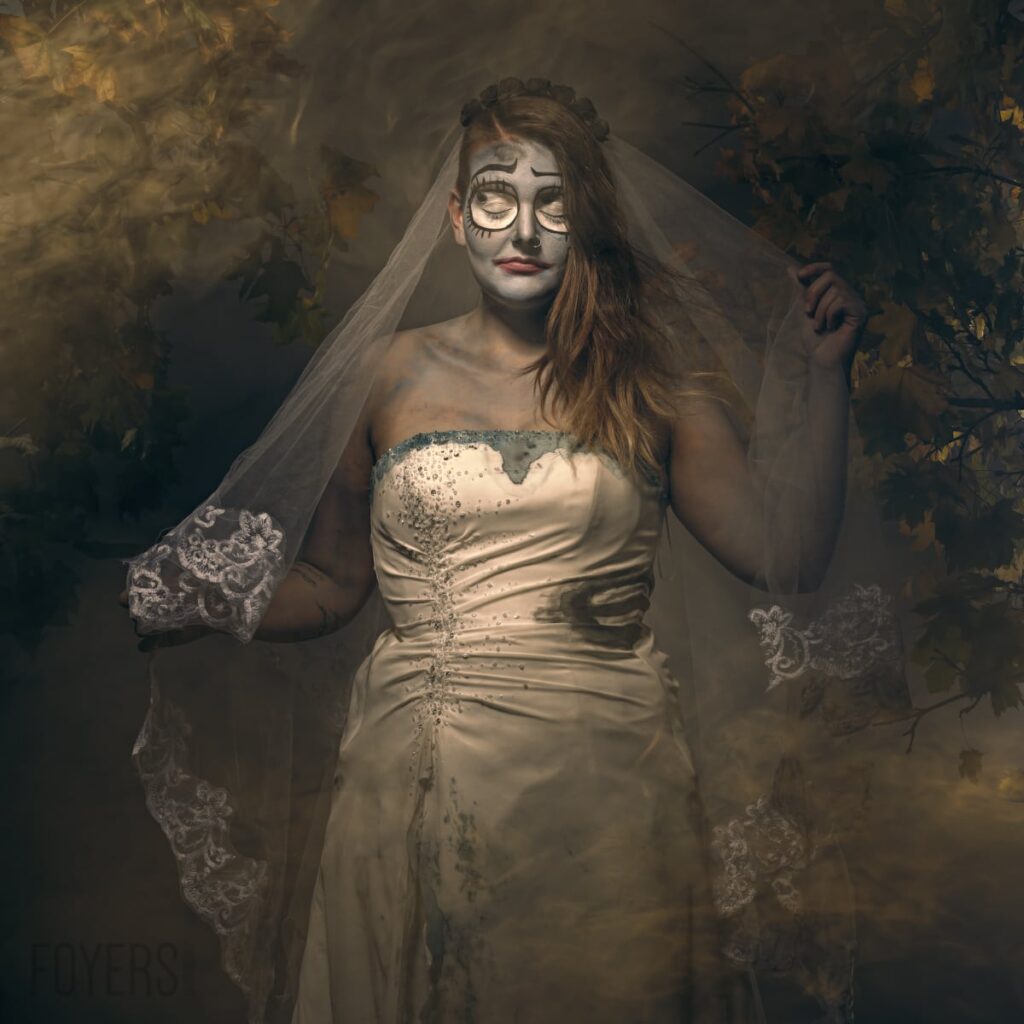 A woman in a white embellished wedding dress wears skeletal face paint and a veil, posed against a dark, textured golden-brown background.