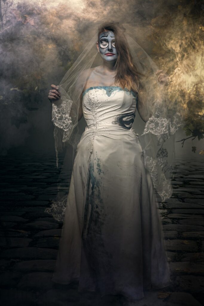 A woman in a white wedding dress with skeletal face paint and a lace veil stands in a misty, atmospheric setting with dark wooden flooring.
