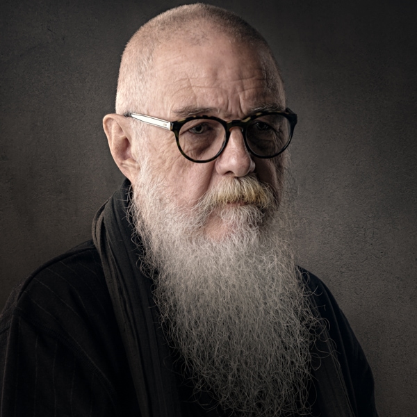 An elderly man with a long white beard wearing glasses and dark clothing, photographed against a grey background.