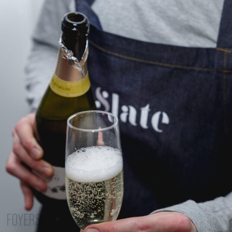 A hand holds a glass of sparkling wine next to a bottle. The person wears a dark apron with partial text visible.
