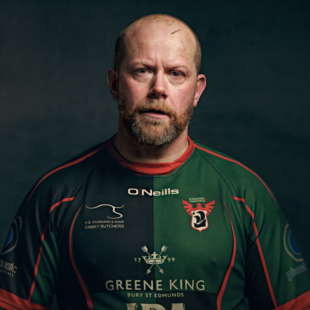 A man with a beard wearing a green and black rugby shirt with red trim, photographed against a dark teal background.