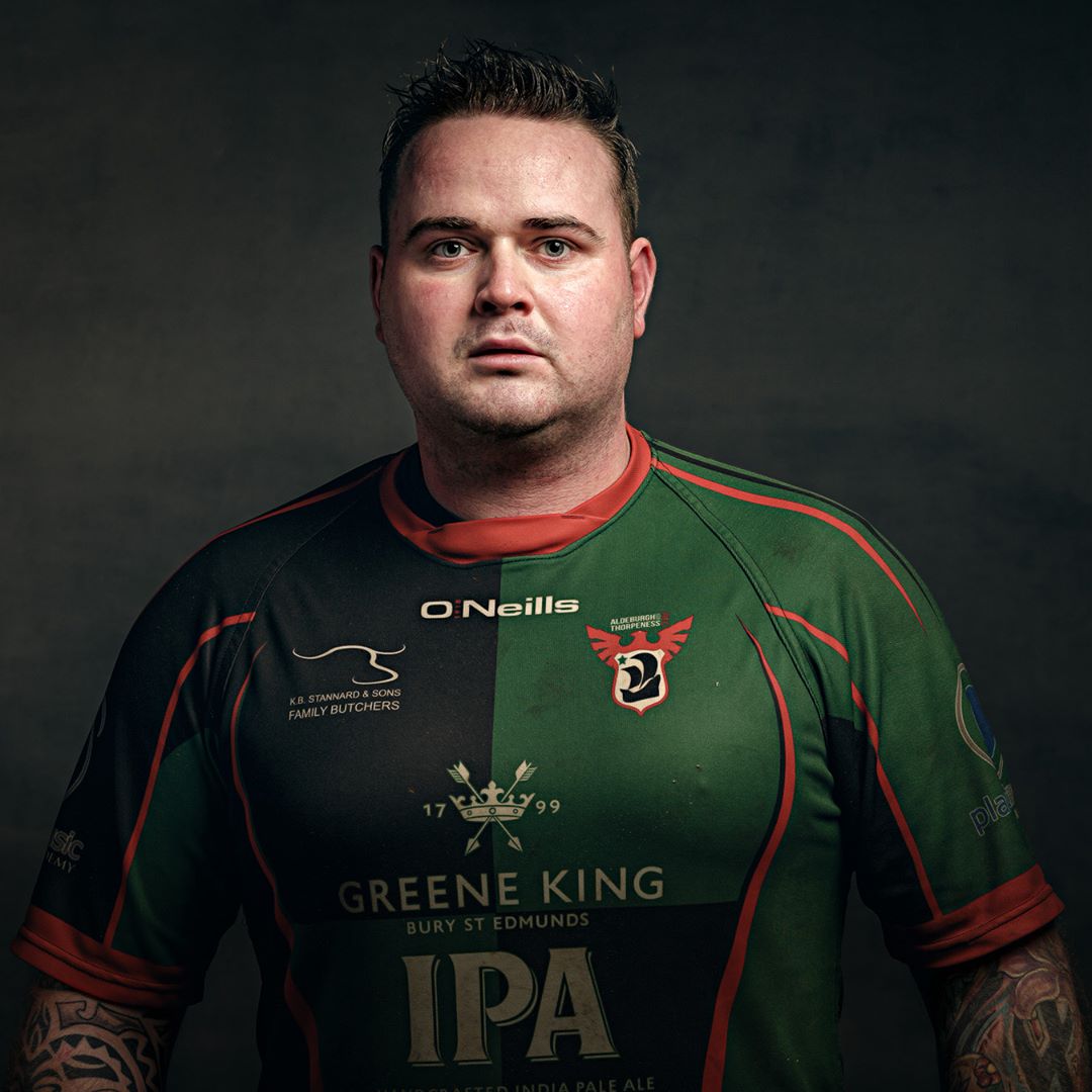 A man wearing a green and black rugby jersey with red trim, photographed against a dark background in a professional portrait style.
