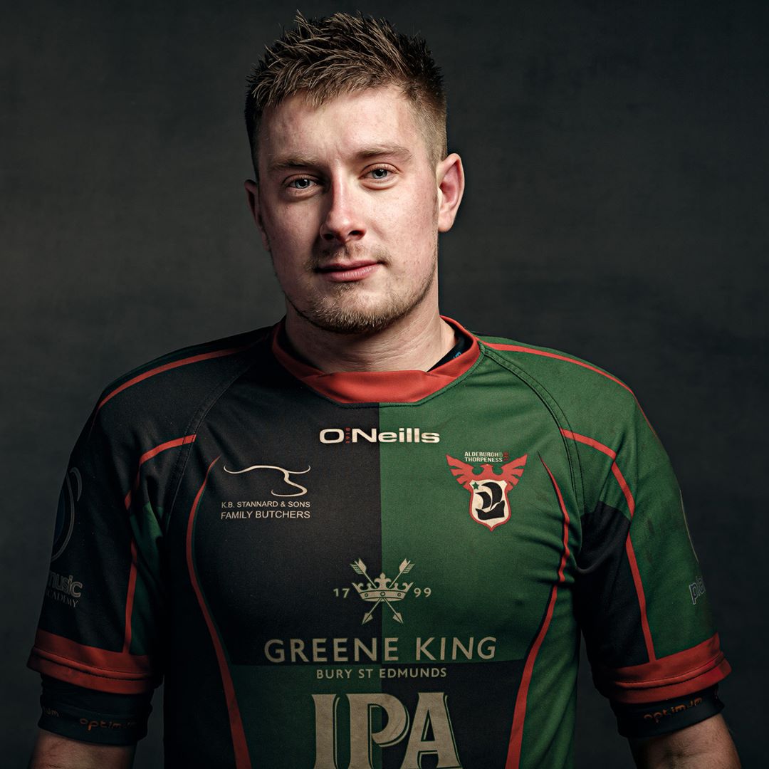 A man wearing a green and black rugby jersey with red trim, photographed against a dark grey background.