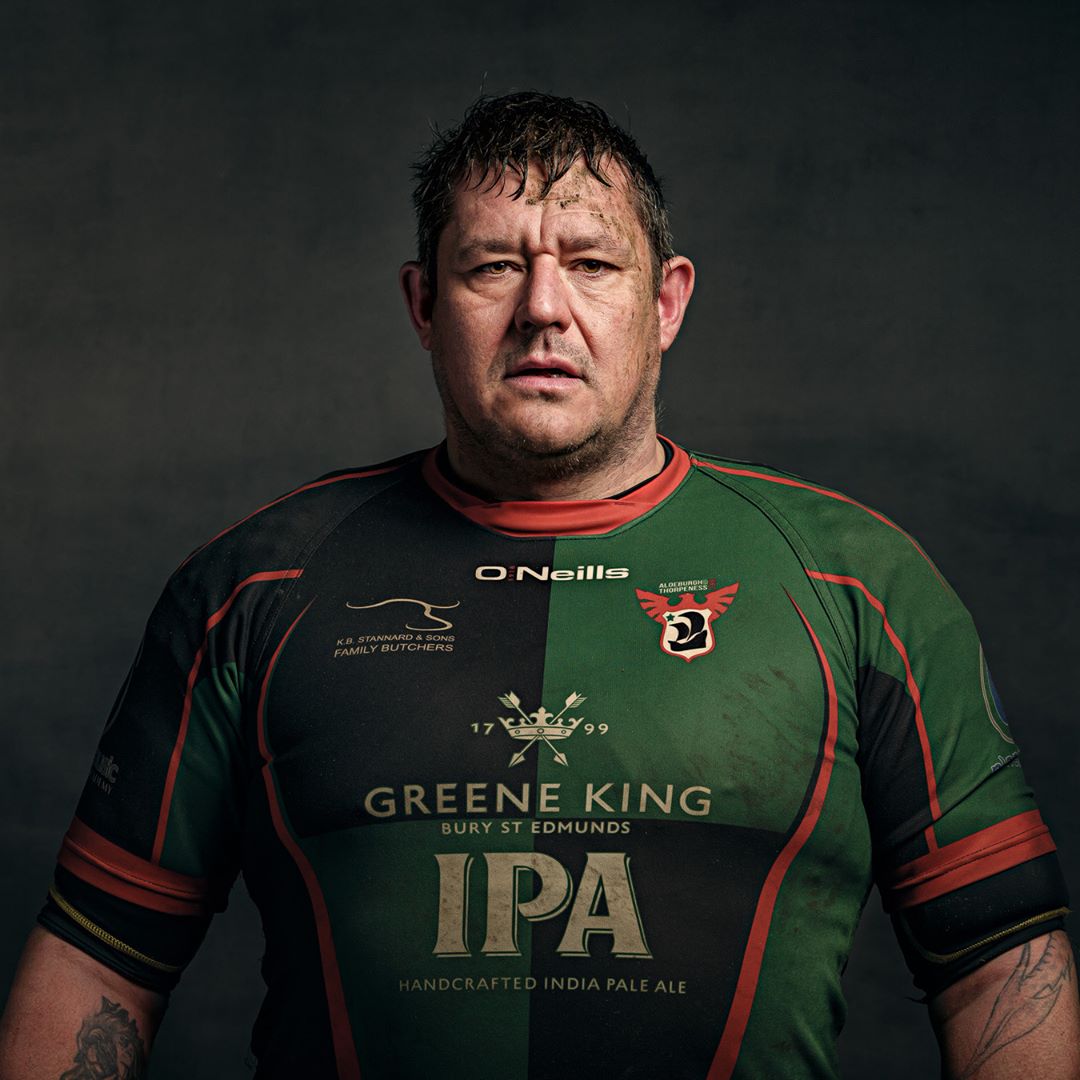 A man wearing a rugby jersey with green and black panels and red trim, featuring sponsor logos and Greene King IPA branding, photographed against a dark grey background.
