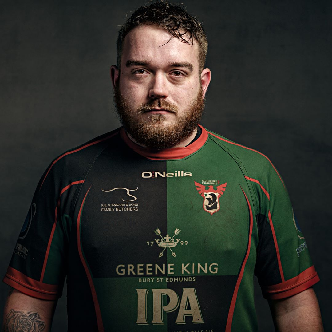 A man with a full beard wearing a green and black rugby jersey with red trim, photographed against a dark background.