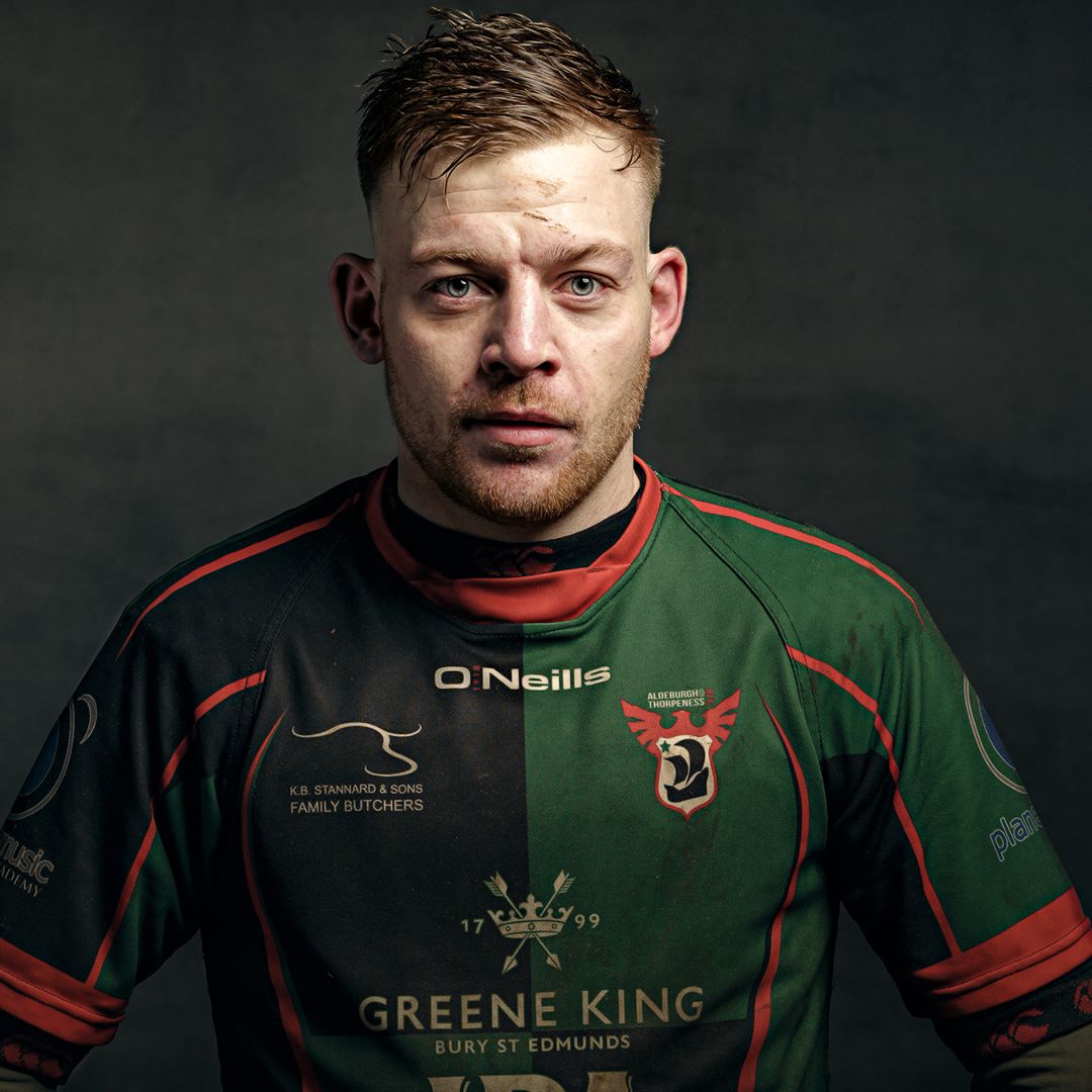 Man wearing green and black rugby jersey with red trim, photographed against dark grey background. Professional portrait style.
