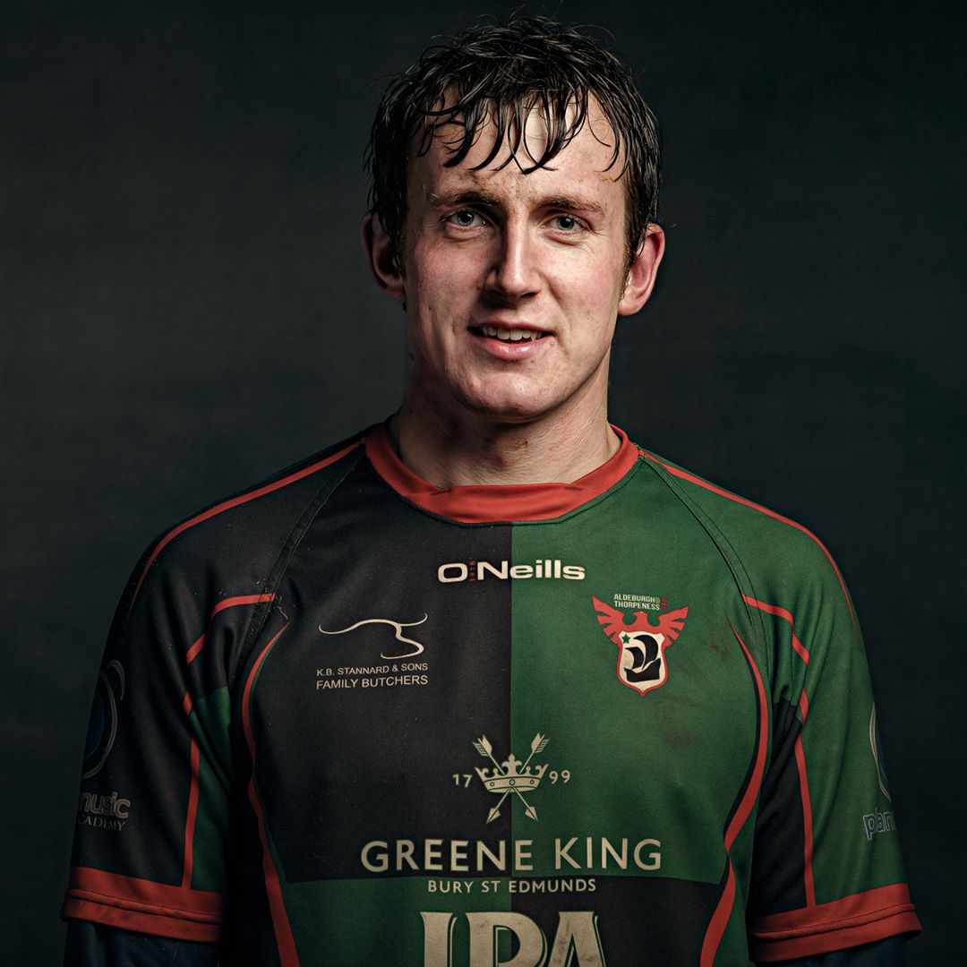 A man in a green and black rugby jersey with red trim, smiling at the camera against a dark background.