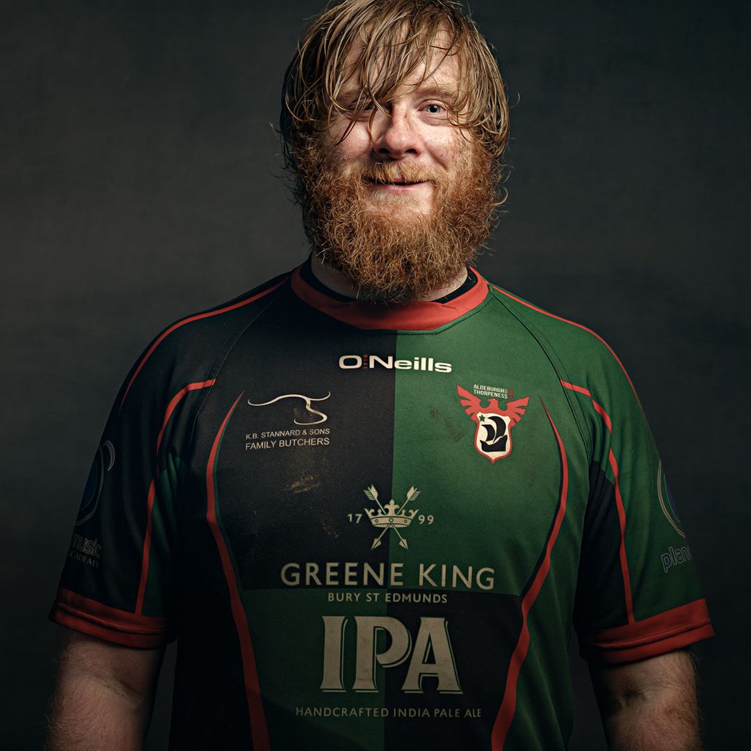 A man with a full beard and shoulder-length brown hair wears a green and black rugby shirt with red trim against a dark background.