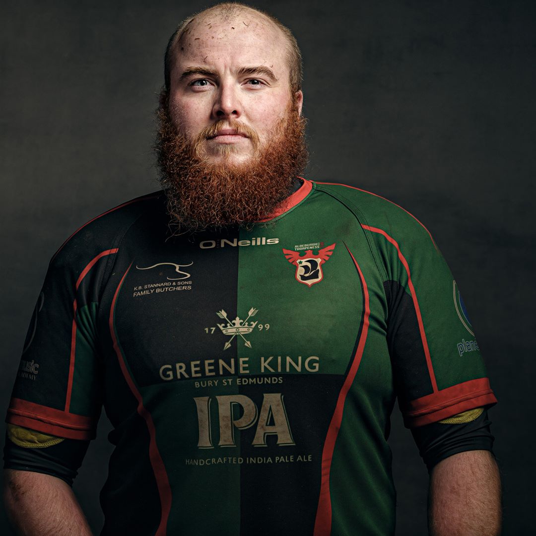 A man with a long reddish-brown beard wearing a green and black rugby jersey with red trim, sponsored by Greene King IPA.