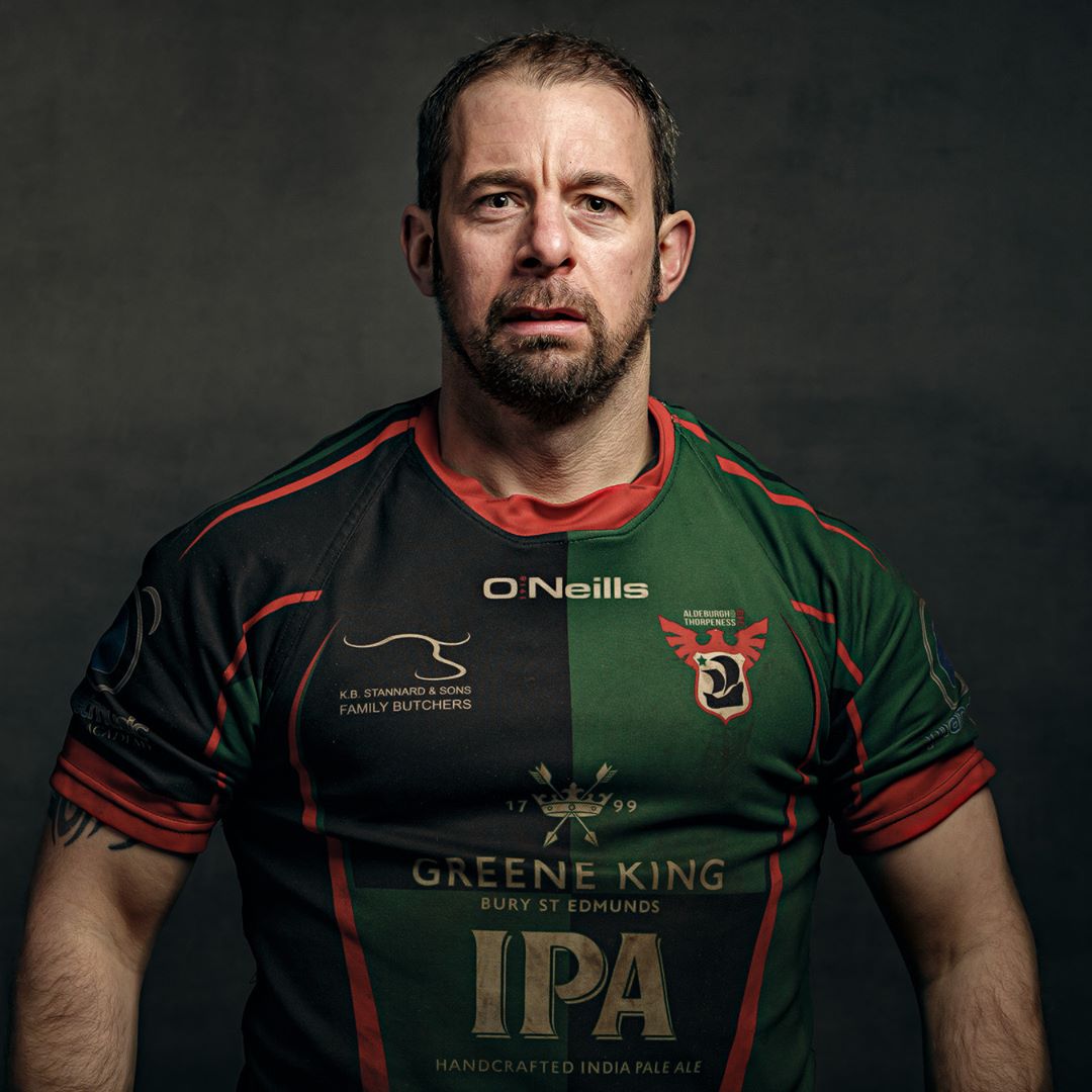 A man wearing a green and red rugby jersey with sponsor logos poses for a portrait against a dark grey background.